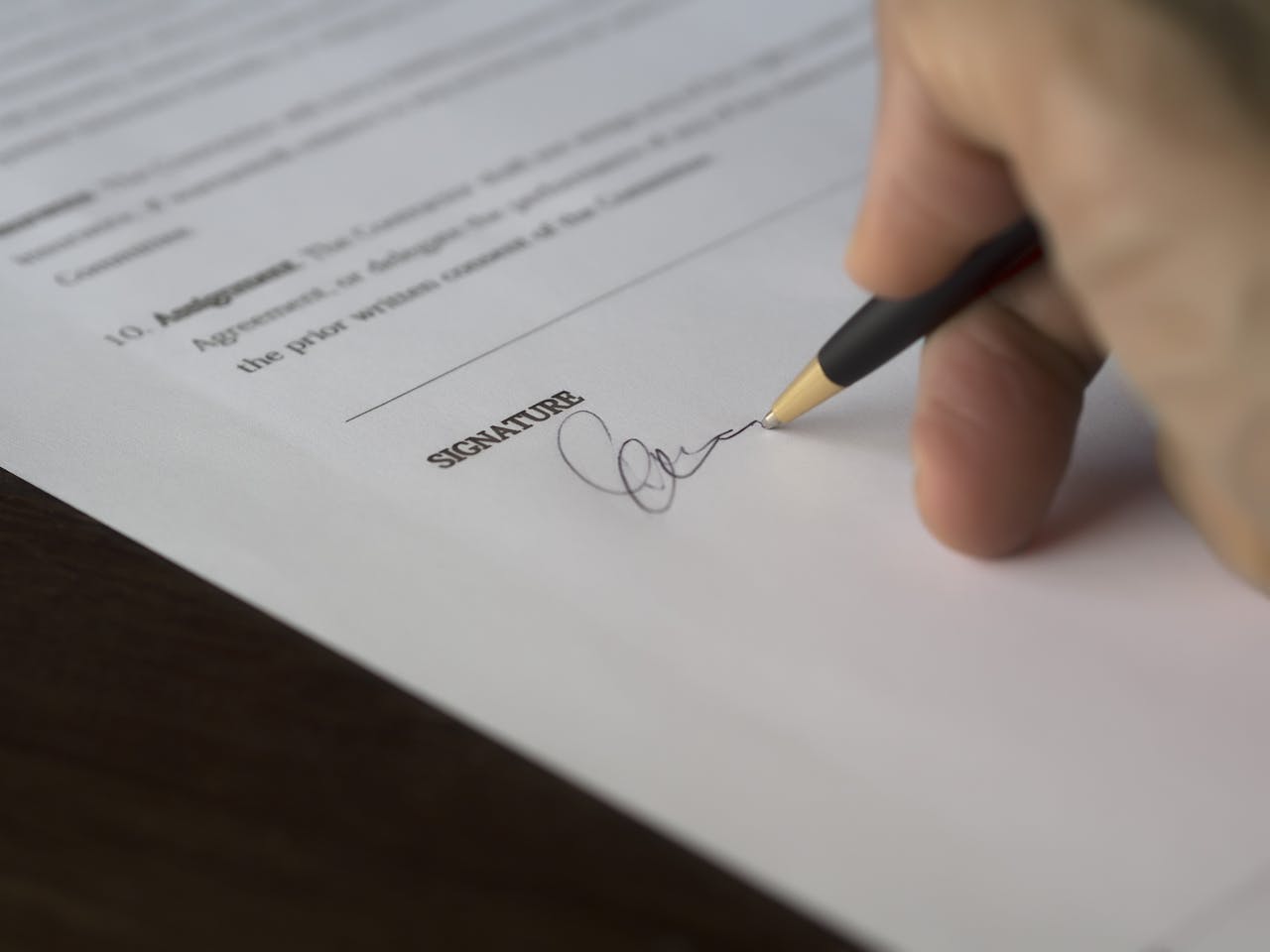 A hand signs a formal contract with a pen on a wooden desk.